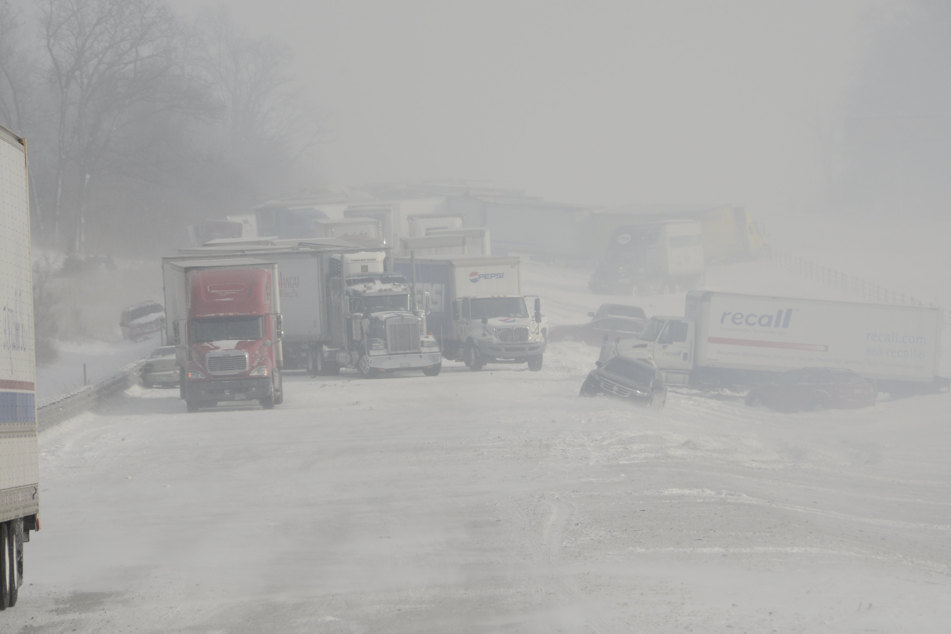 A snow squall limits visibility along a highway.  In the distance, semi trucks and cars can be seen strewn along the road, with some damaged.