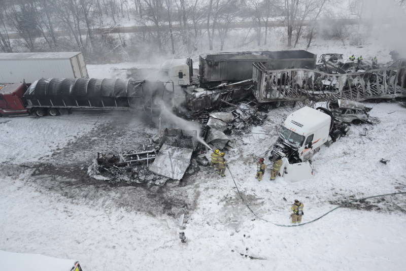 Firefighters spray water onto the burned out wreckage of several semi trucks amidst snow covered vehicles scattered across the highway and its shoulders.